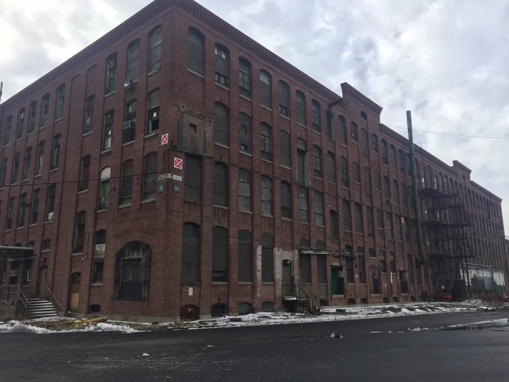 A large, old brick industrial building with many windows, most of them boarded up. Snow is scattered on the ground and the street is empty under a cloudy sky.